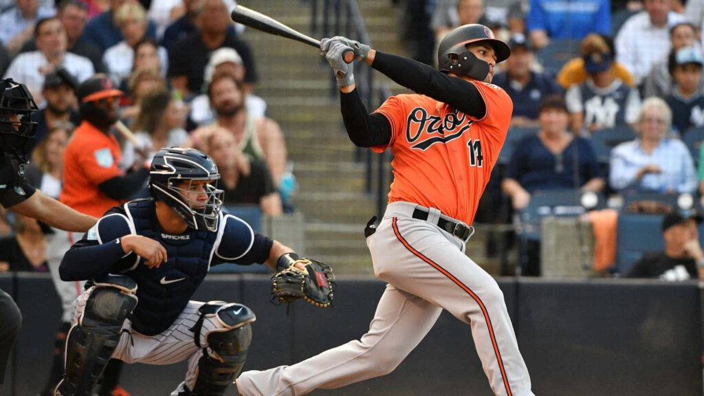Austin Hays swings bat in Orioles uniform during baseball game with players and fans background Wallpaper