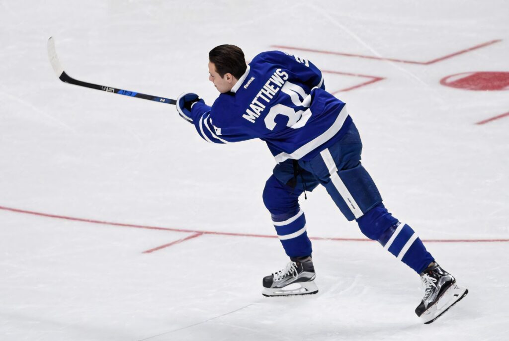 Auston Matthews shooting puck on ice in blue Toronto Maple Leafs jersey with rink red lines background Wallpaper