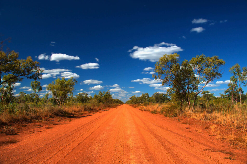 Australian Outback dirt road under bright blue sky bordered by native trees and dry grasses Wallpaper
