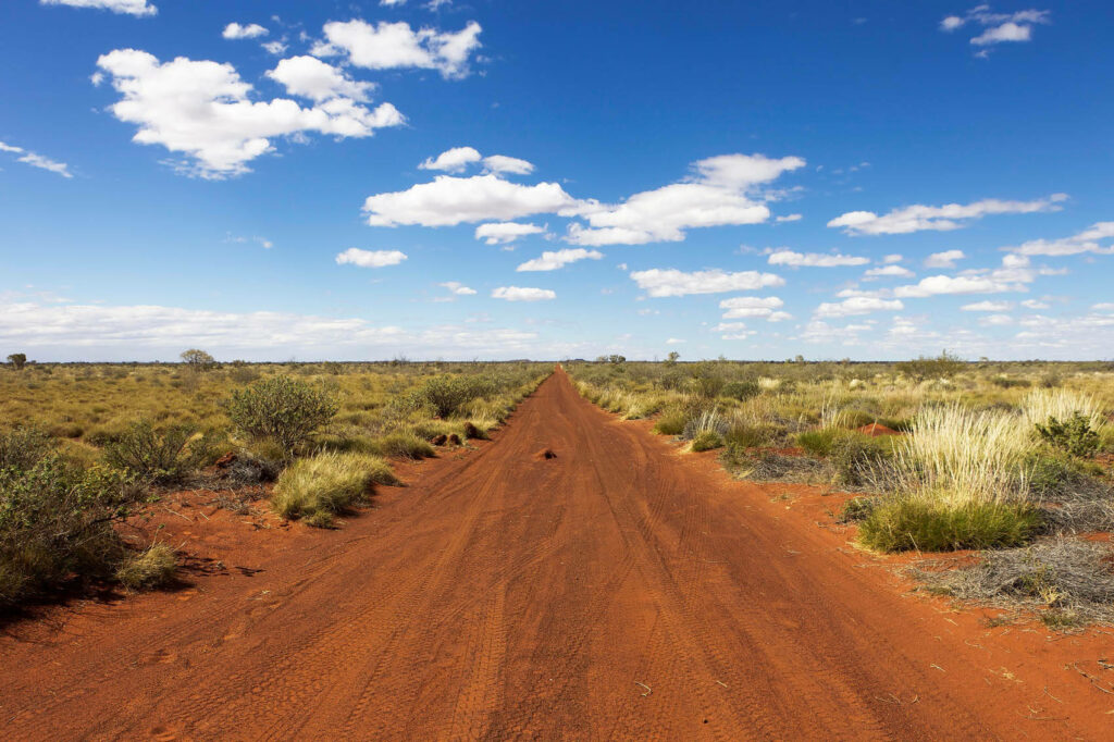 Red dirt car tracks in Australian Outback under blue sky with white clouds and native shrubs Wallpaper