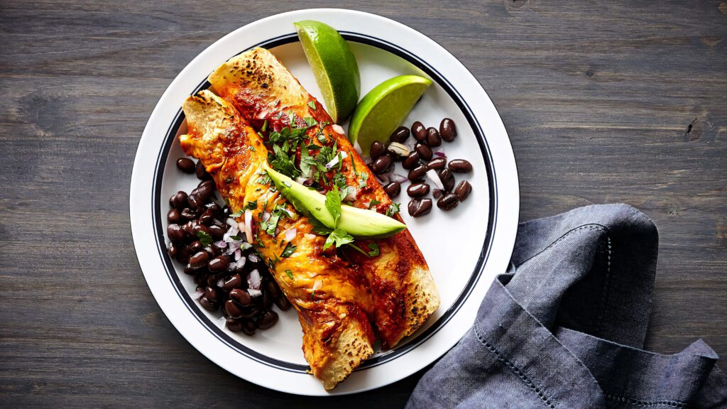 Plate of enchiladas with avocado cilantro black beans and lime wedges on rustic wooden table Wallpaper