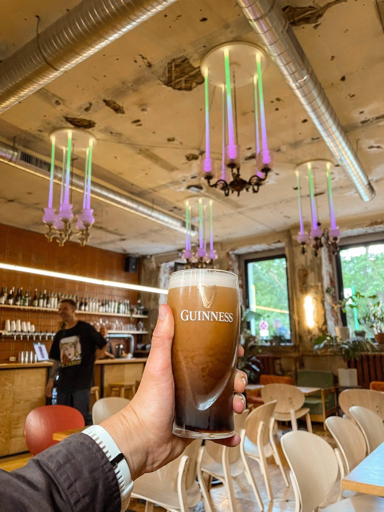 Hand holding a fresh Guinness Irish Dry Stout pint in a rustic bar with neon chandeliers wallpaper