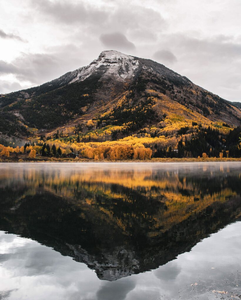 Mountain peak with autumn yellow trees reflected in serene lake under cloudy sky wallpaper background