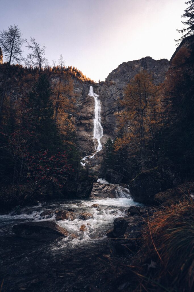 Waterfall cascading down rugged cliff into river surrounded by golden autumn trees at sunset Wallpaper