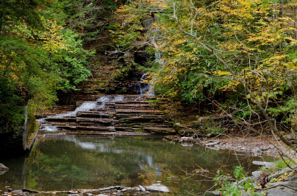 Serene waterfall over rocks with autumn foliage and calm water reflecting a peaceful forest scene Wallpaper
