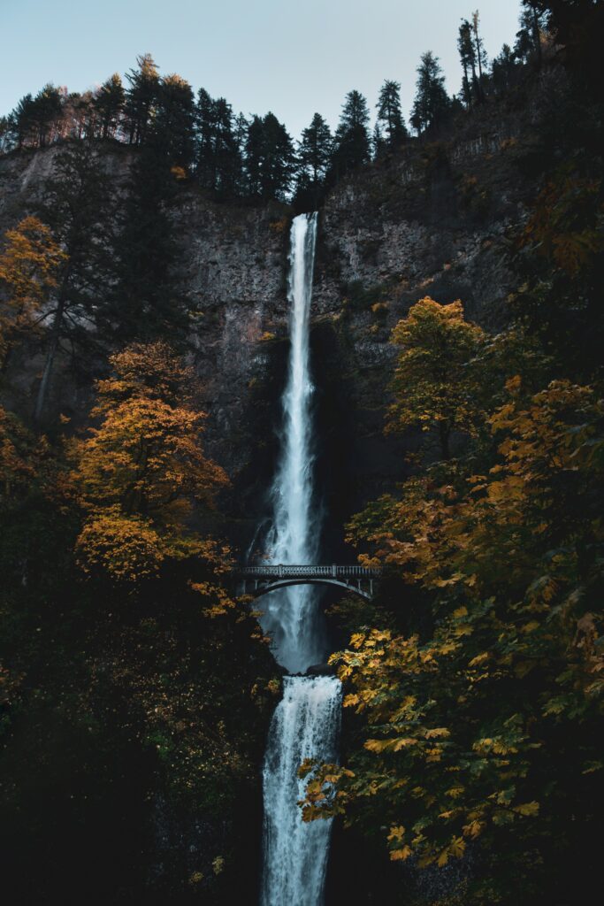 Tall waterfall over rugged cliff with autumn trees and charming bridge serene nature wallpaper