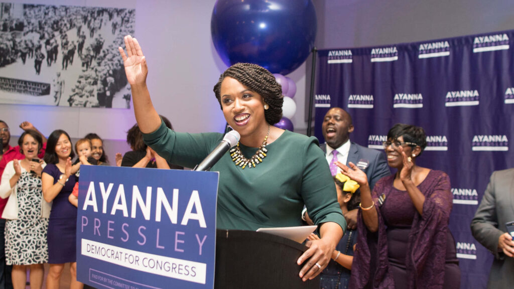 Ayanna Pressley waves to supporters at a lively campaign event with cheering attendees and banners Wallpaper