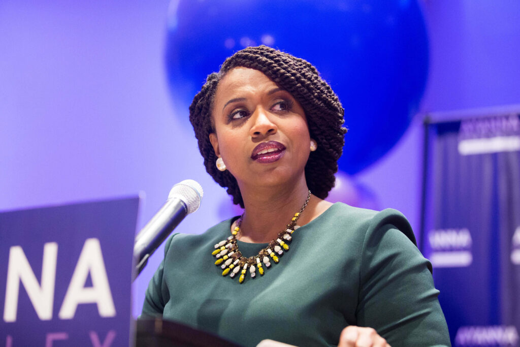 Representative Ayanna Pressley speaking confidently at podium with vivid violet wallpaper background
