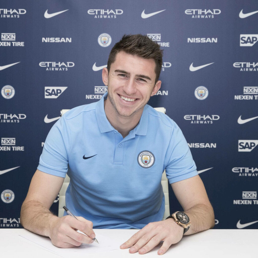 Aymeric Laporte smiling signing contract at table with Manchester City backdrop wallpaper background