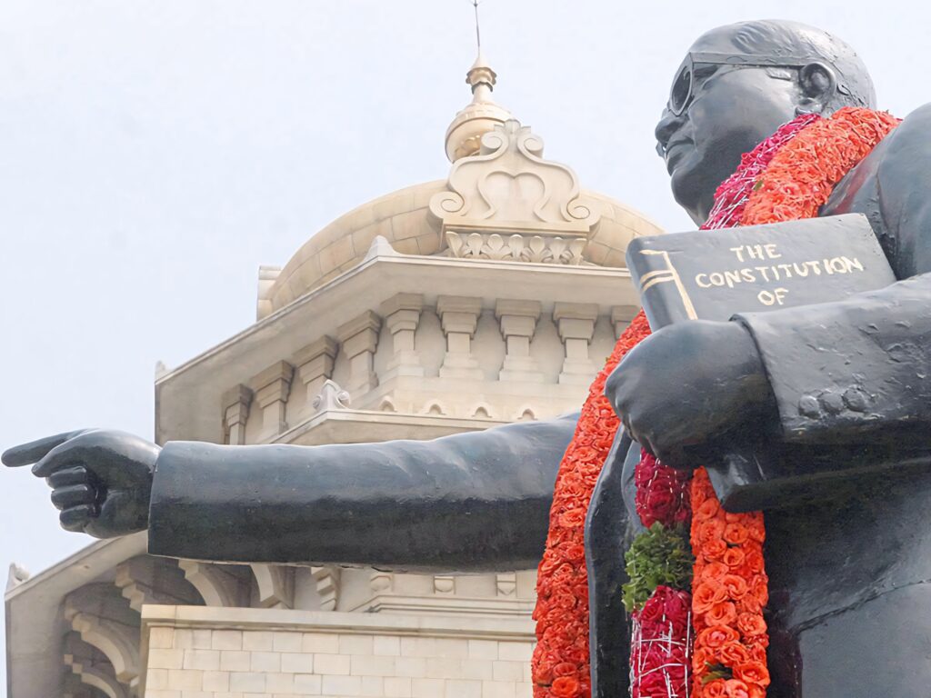 Statue of B R Ambedkar with flower garlands holding Indian Constitution against architecture background Wallpaper