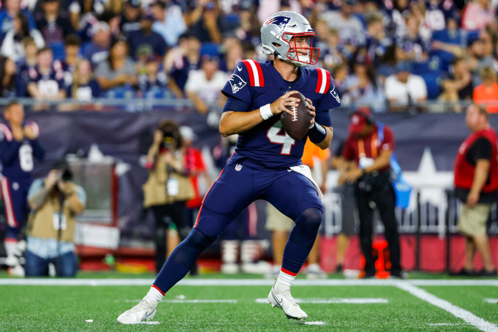 Bailey Zappe throwing football in New England Patriots uniform mid play on field wallpaper