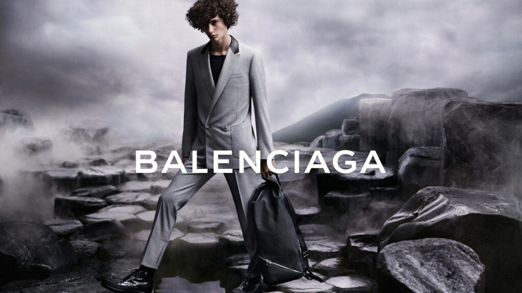 Balenciaga male model in gray suit on rugged rocks with black backpack under moody sky wallpaper
