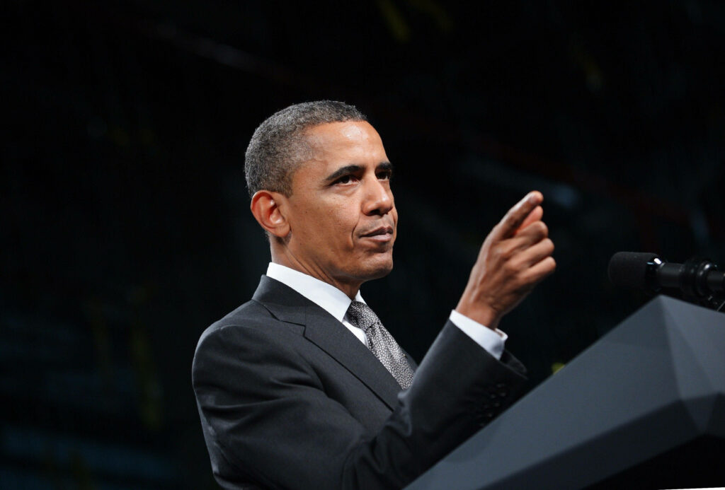 Barack Obama in sharp suit delivering speech confidently pointing hand at podium with wallpaper background