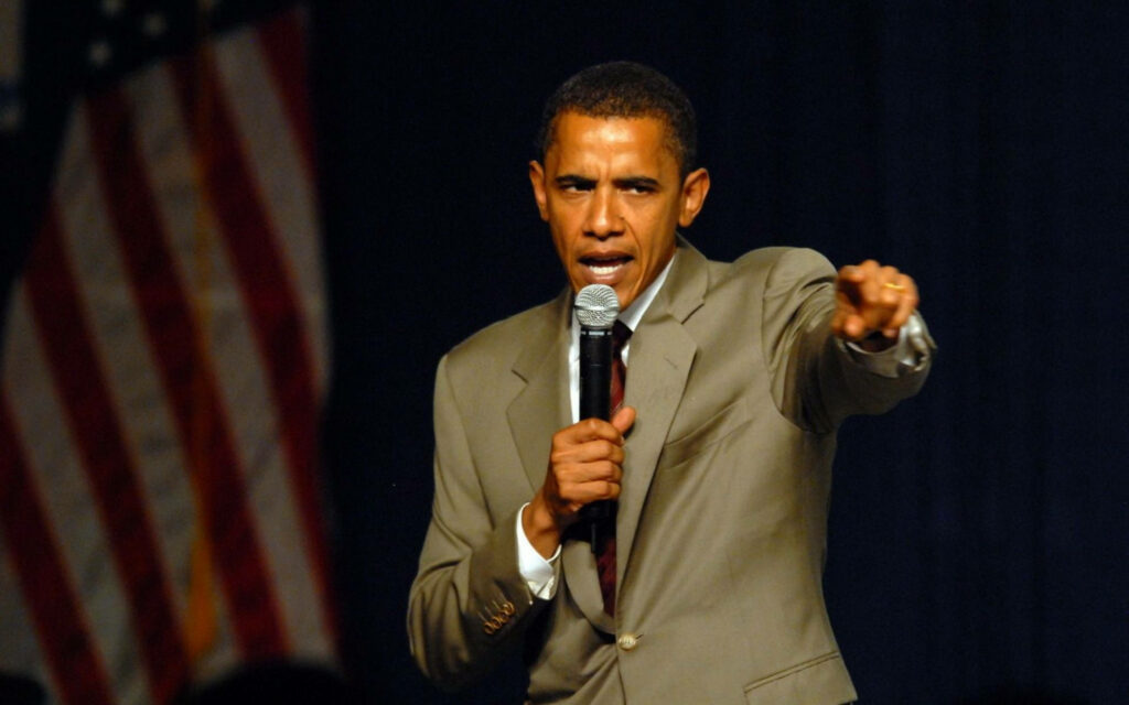 Barack Obama delivering passionate speech holding microphone pointing forward with US flag background Wallpaper