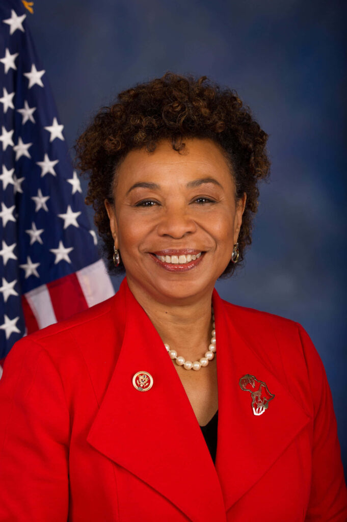 Barbara Lee in red blazer with pearl necklace standing confidently before American flag background Wallpaper