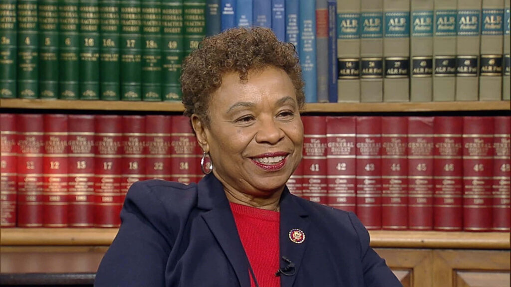 Barbara Lee smiling seated in colorful library surrounded by bookshelves scholarly warm setting Wallpaper