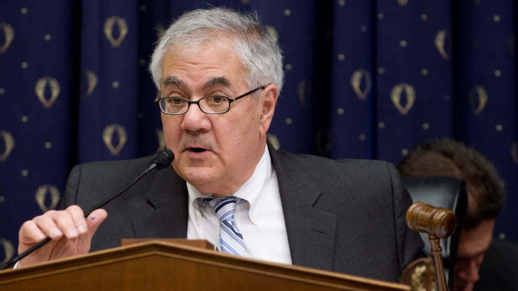 Barney Frank speaking into microphone in suit and tie with dark patterned wallpaper background