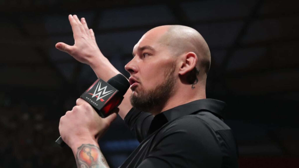 Wrestler Baron Corbin speaking passionately in WWE ring holding microphone with wallpaper background