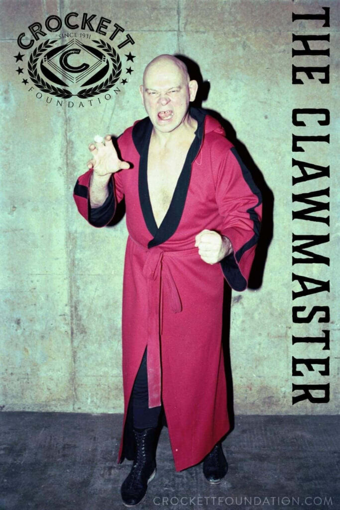 Baron Von Raschke in red robe posing with claw hold wrestling move against concrete wall background Wallpaper