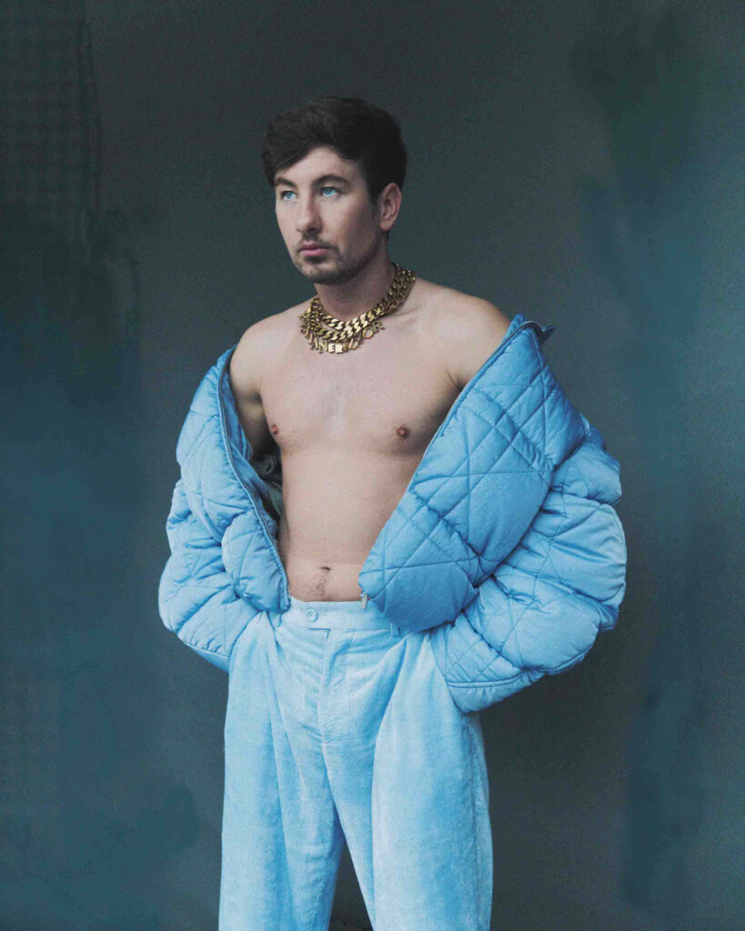 Barry Keoghan in stylish blue puffer jacket with gold necklace on minimal wallpaper background