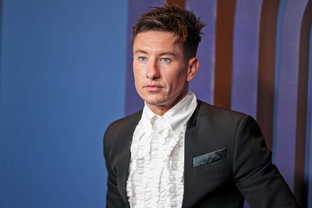 Barry Keoghan in sharp suit with ruffled shirt at stylish event against modern wallpaper background