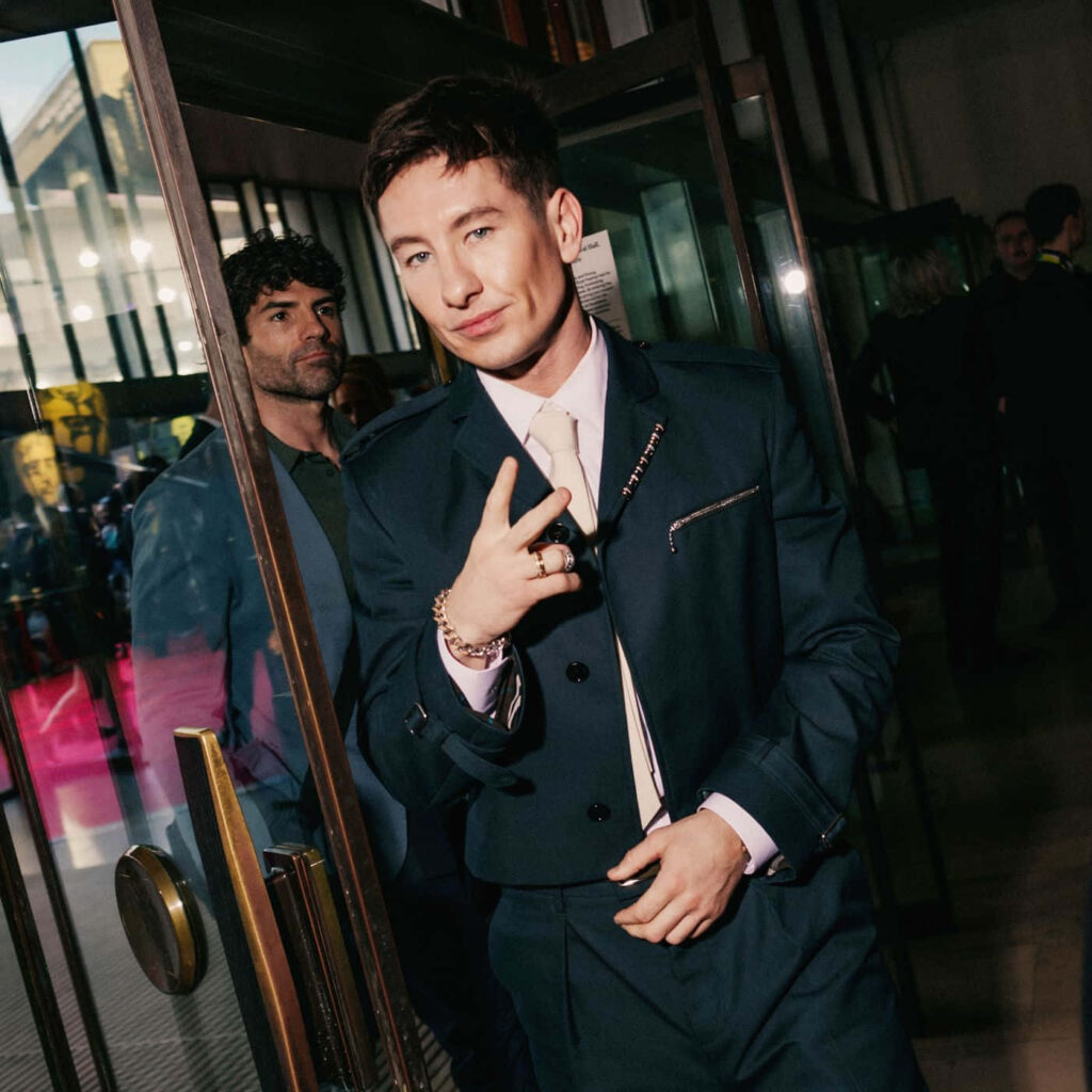 Barry Keoghan in sharp suit making peace sign exiting event through glass doors with wallpaper background