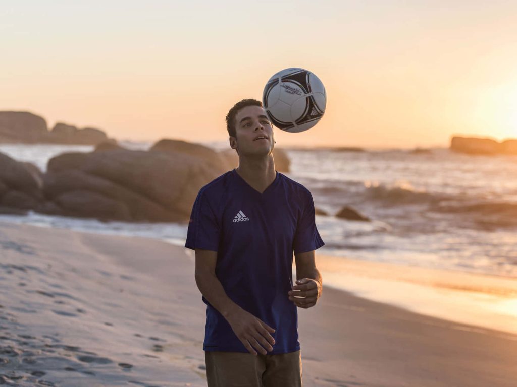 Young man in blue Adidas shirt balances soccer ball on forehead at sunset beach freestyle Wallpaper