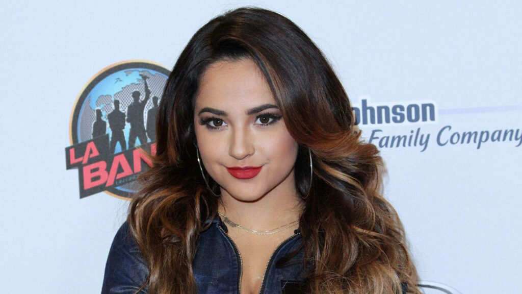 Becky G at La Banda event with voluminous hair bold red lipstick and branded wallpaper background