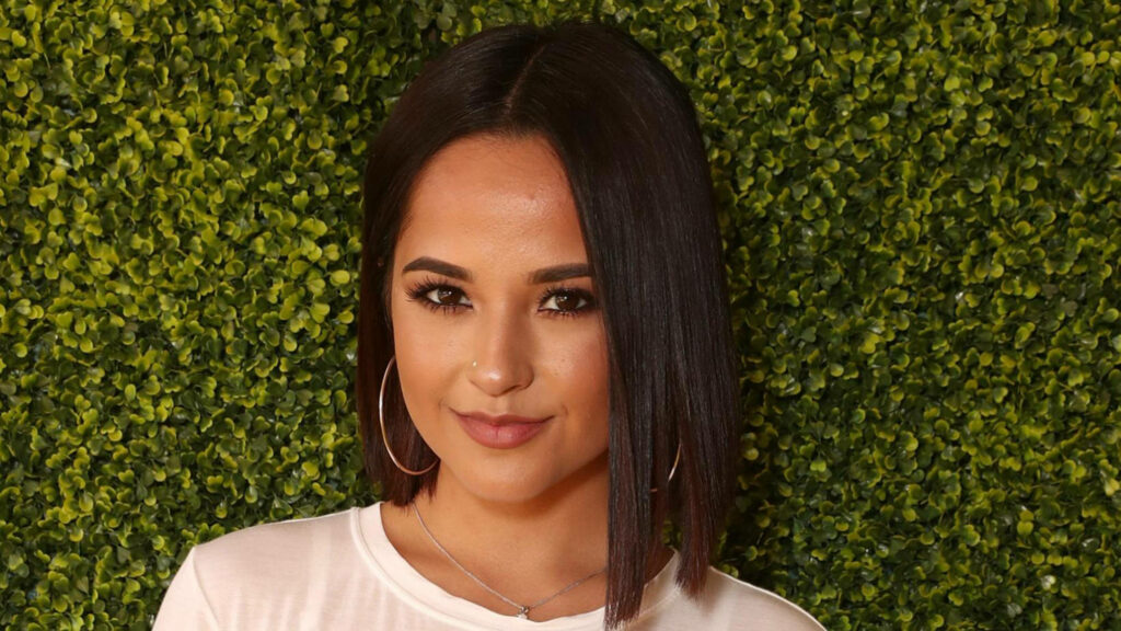 Becky G confidently posing in front of lush green plants at Despicable Me event wallpaper background
