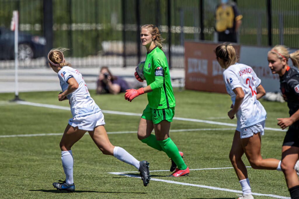 Bella Bixby in bright green goalkeeper kit focused during soccer match with players in action Wallpaper