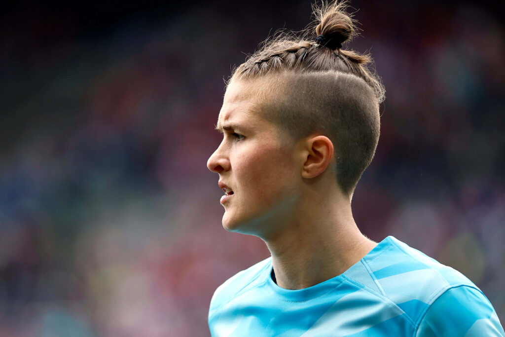 Bella Bixby goalkeeper profile with distinctive hairstyle in kit against blurred stadium background Wallpaper