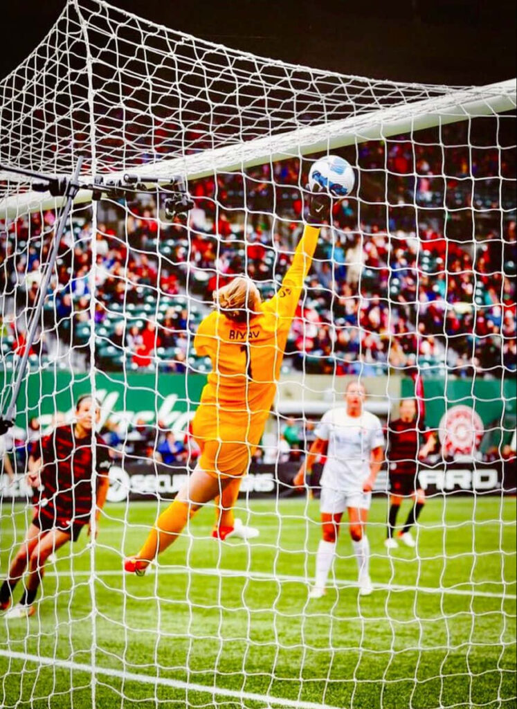 Bella Bixby airborne making a stunning soccer save in intense match with fans in stadium background Wallpaper