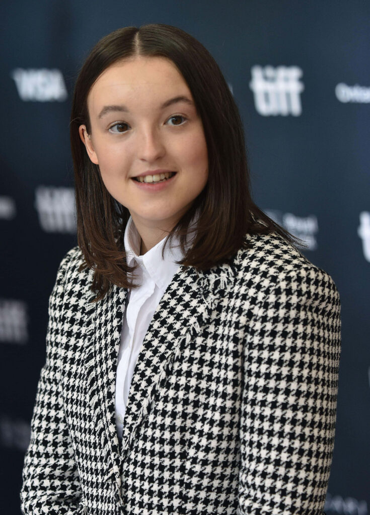 Bella Ramsey in stylish houndstooth jacket and white shirt posing dark backdrop wallpaper