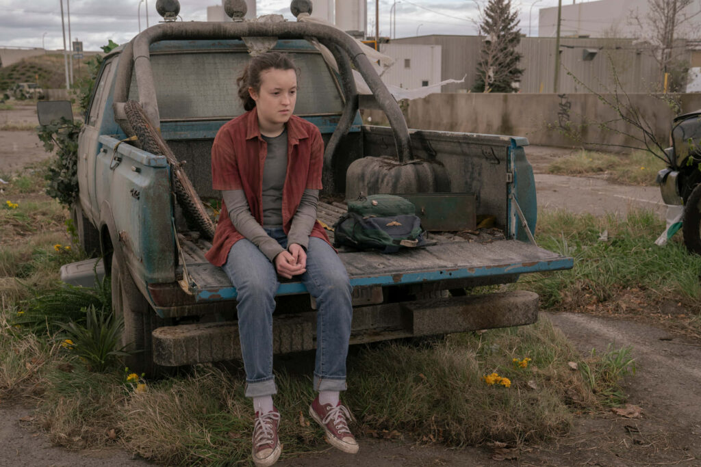 Bella Ramsey sitting on rusty truck tailgate in overgrown abandoned post-apocalyptic setting wallpaper