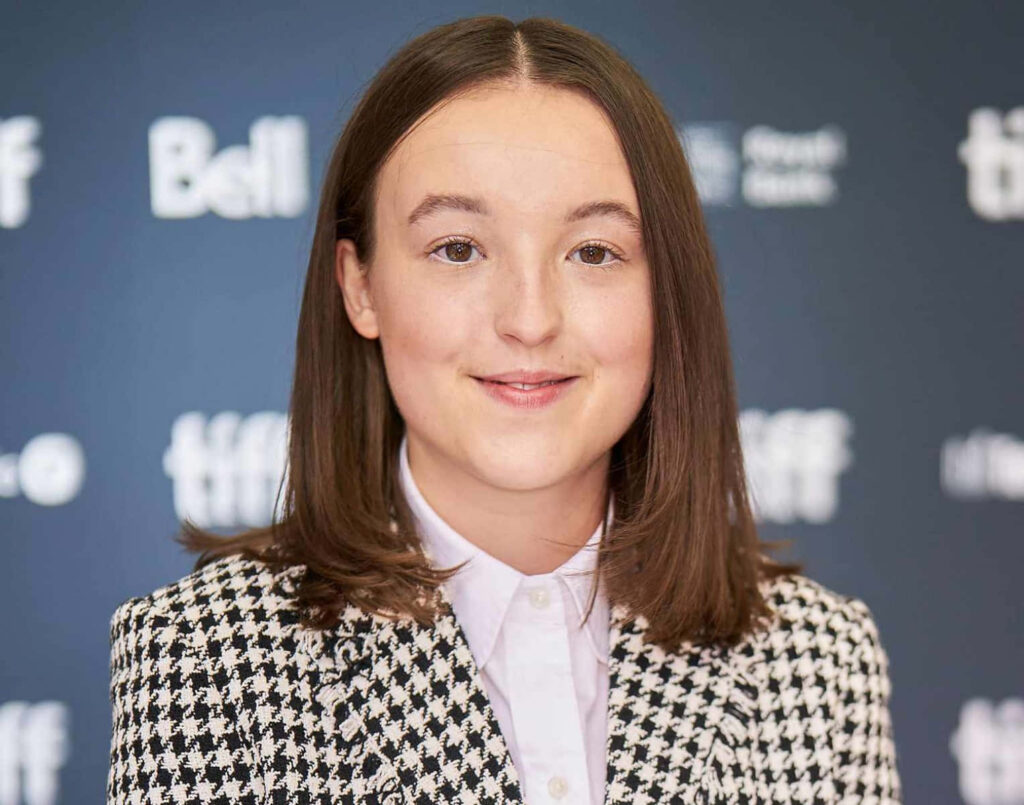 Bella Ramsey in stylish houndstooth blazer and white shirt at event with logo backdrop wallpaper