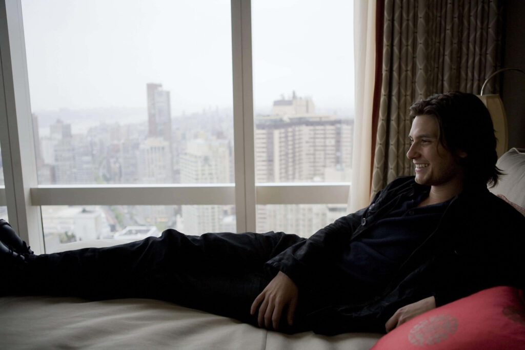Ben Barnes relaxing by window with cityscape and tall buildings in scenic portrait wallpaper