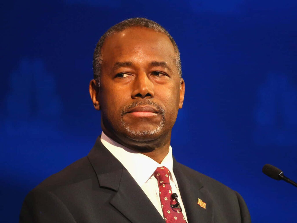 Ben Carson in formal suit and red tie with calm serious expression deep blue background wallpaper
