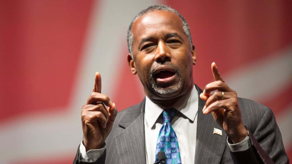 Ben Carson speaking passionately with expressive hand gestures on a professional wallpaper background