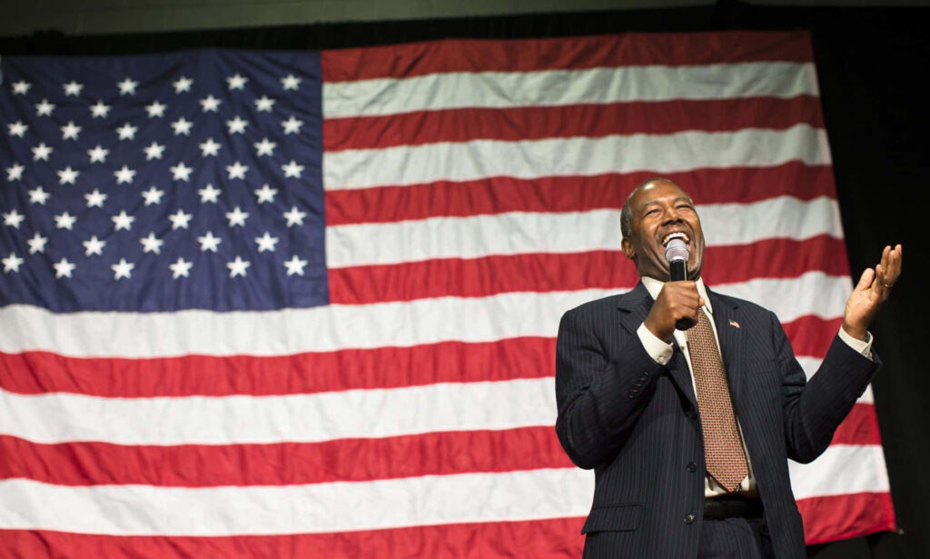 Ben Carson laughing on stage with large American flag background patriotic wallpaper