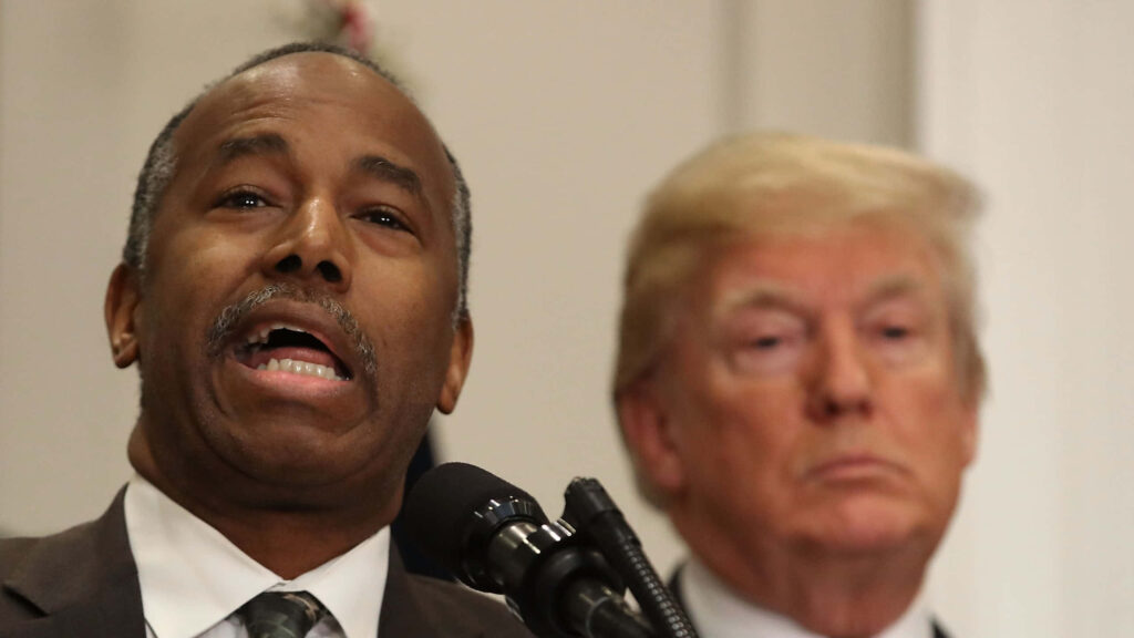 Ben Carson speaking at podium with Donald Trump in background during presidential campaign event wallpaper