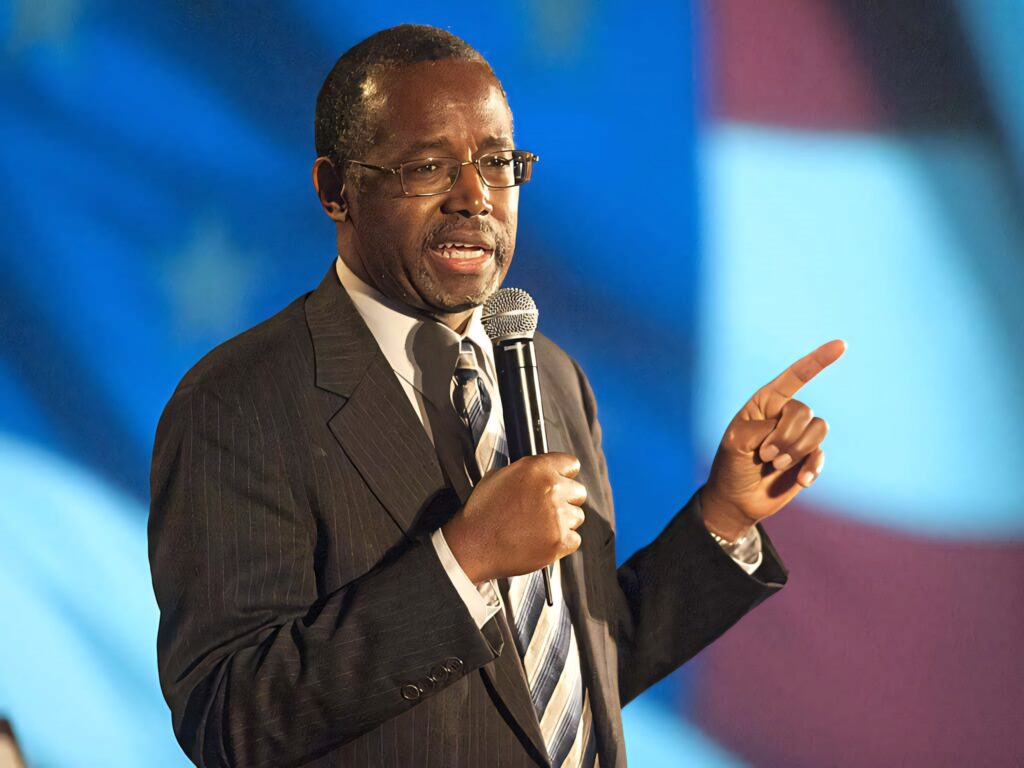 Ben Carson speaking at public event holding microphone with American flag wallpaper background