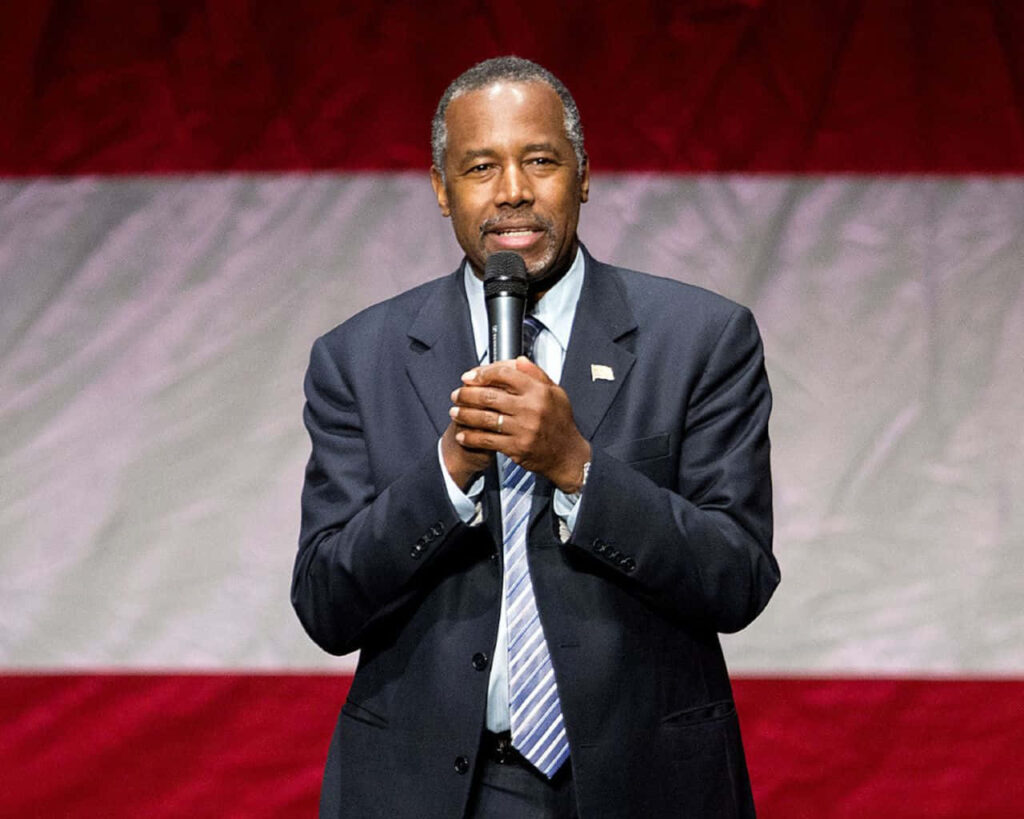 Ben Carson speaking confidently in formal suit with microphone American flag background wallpaper