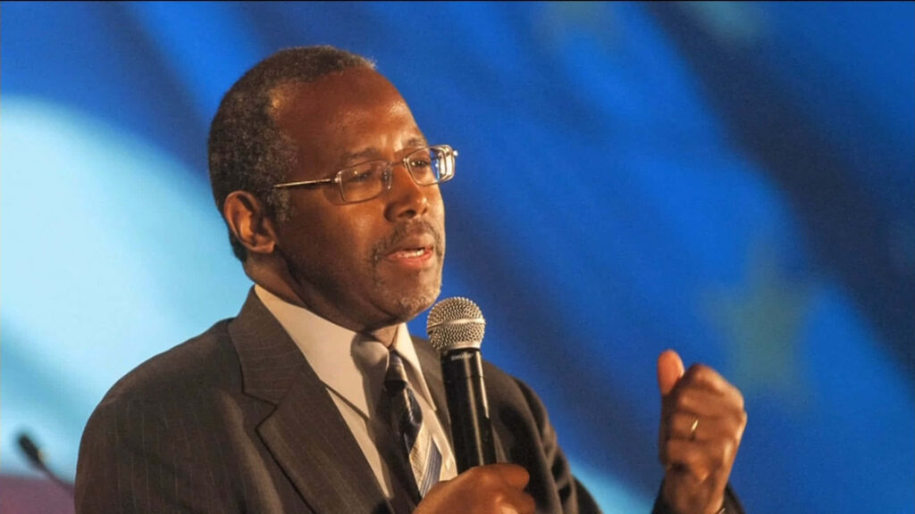 Ben Carson speaking passionately with microphone in suit against blue wallpaper background