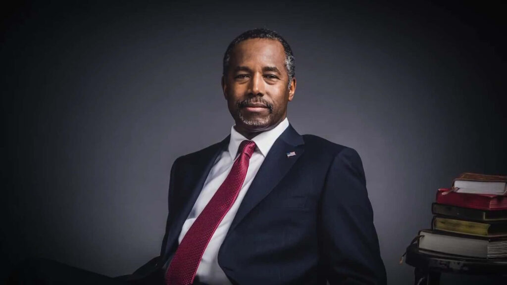 Thoughtful Ben Carson in suit with red tie and his inspiring books stacked beside him wallpaper