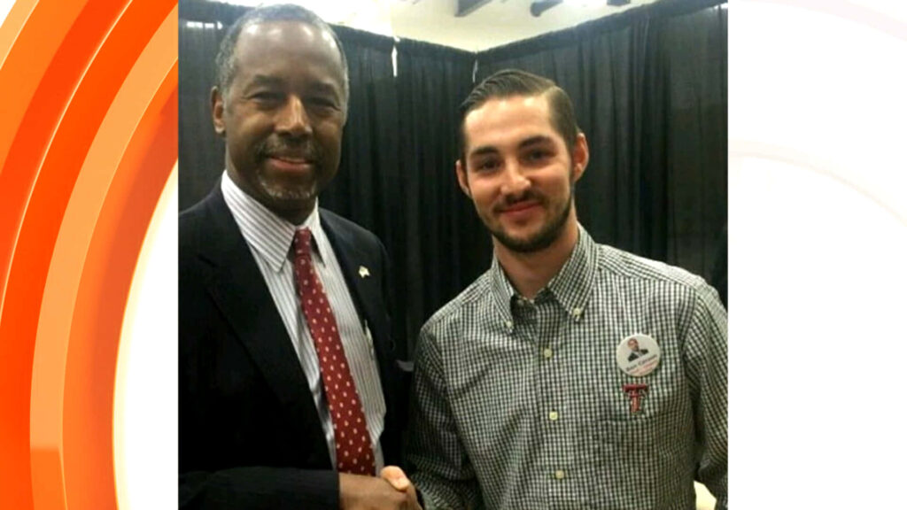 Ben Carson and Braden Joplin smiling and shaking hands in formal event with dark curtain backdrop Wallpaper