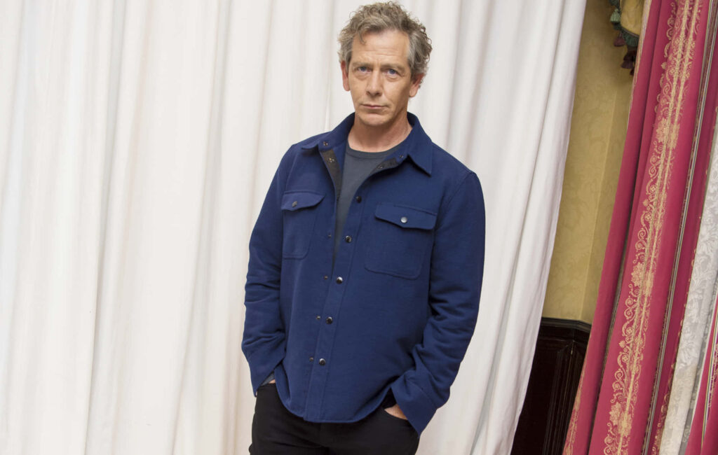 Ben Mendelsohn in stylish blue jacket standing confidently with hands in pockets white curtains background Wallpaper