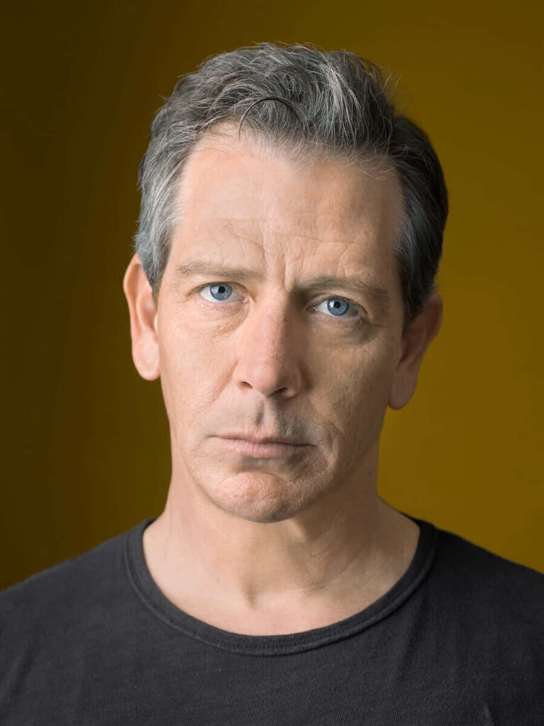 Striking portrait of Ben Mendelsohn with intense gaze on rich gold minimalist wallpaper background