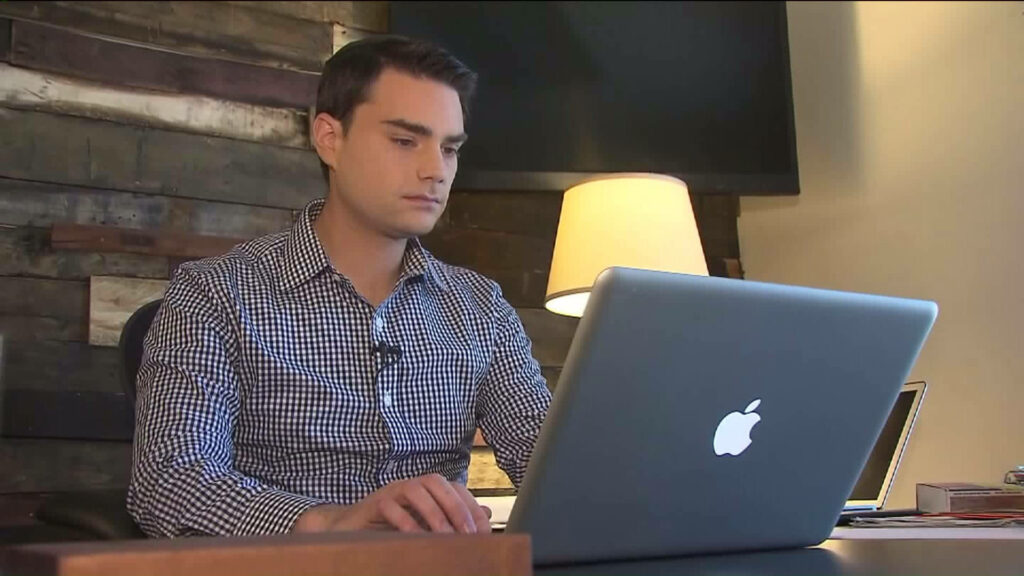 Ben Shapiro focused working on laptop in modern warm lit workspace with wallpaper background