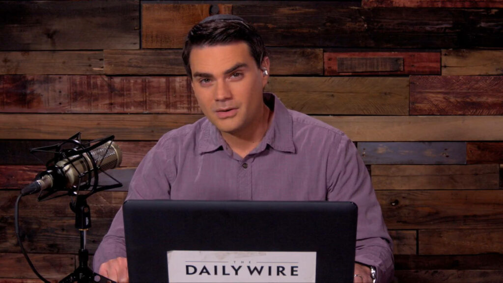 Ben Shapiro hosting show at desk with microphone laptop rustic wooden studio backdrop wallpaper