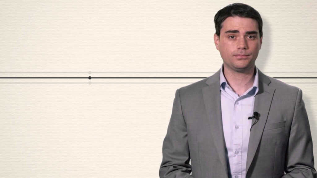 Ben Shapiro in suit speaking confidently in professional setting with light wallpaper background